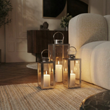 Trois lanternes en métal brillant argenté avec anse arrondie, dessus plat et avec fenêtres transparentes en verre. Lanternes posées au sol dans un salon moderne à côté d'un canapé blanc. Ambiance tamisée. Trois tailles différentes.