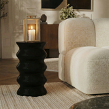 Lanterne en métal mat effet brossé doré avec anse carrée, dessus carré et plat. Avec fenêtres transparentes en verre. Structure dorée en acier inoxydable. Une bougie blanche est allumée à l'intérieur. Lanterne posée sur une table d'appoint ondulée, dans un salon moderne à côté d'un canapé blanc.