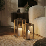 Deux lanternes en métal mat effet brossé doré avec anse carrée, dessus carré et plat. Avec fenêtres transparentes en verre. Structure dorée en acier inoxydable. Une bougie blanche est allumée à l'intérieur de chaque lanterne. Les deux lanternes sont posées au sol sur un tapis, dans un salon moderne à côté d'un canapé blanc.