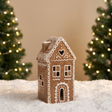 Figurine Chalet de Noël Ginger 16 cm en céramique effet pain d'épices, LED allumée, posée sur la neige devant un sapin lumineux flouté