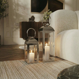 Deux lanternes en métal brillant argenté avec anse arrondie, dessus arrondi et avec fenêtres en transparentes en verre. Lanternes posées au sol dans un salon moderne à côté d'un canapé blanc. Ambiance tamisée. Deux tailles différentes.