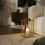 Lanterne en métal mat effet brossé doré avec anse carrée, dessus carré et plat. Avec fenêtres transparentes en verre. Structure dorée en acier inoxydable. Une bougie blanche est allumée à l'intérieur. Lanterne posée au sol, dans un salon moderne à côté d'un canapé blanc.