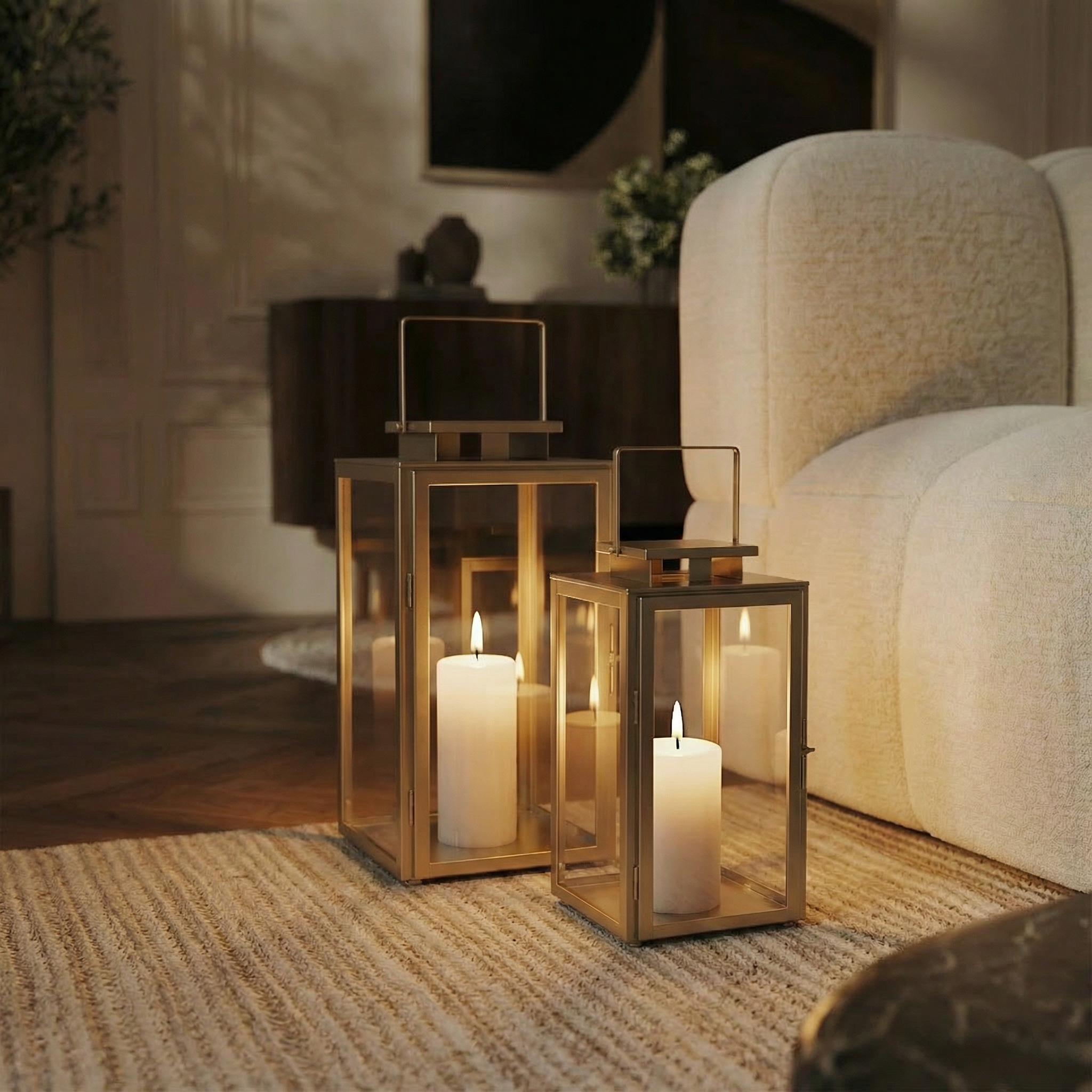 Deux lanternes en métal mat effet brossé doré avec anse carrée, dessus carré et plat. Avec fenêtres transparentes en verre. Structure dorée en acier inoxydable. Une bougie blanche est allumée à l'intérieur de chaque lanterne.  Les deux lanternes sont posées au sol sur un tapis, dans un salon moderne à côté d'un canapé blanc.