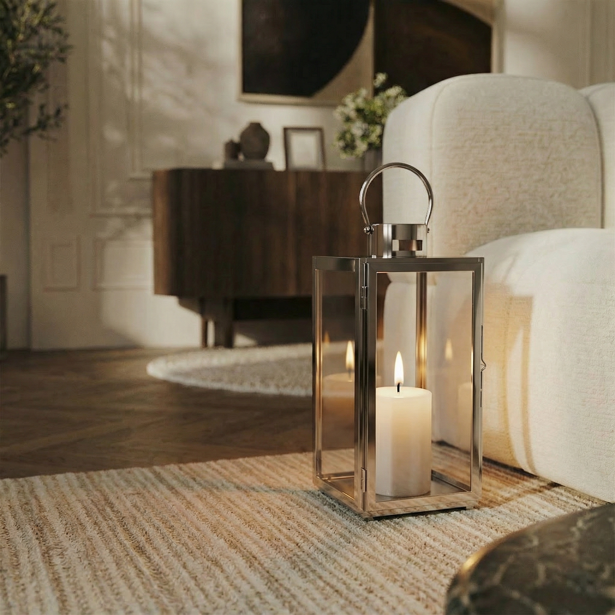 Lanterne en métal brillant argenté avec anse arrondie, dessus plat et avec fenêtres transparentes en verre. Une bougie blanche est allumée à l'intérieur. Lanterne posée au sol, dans un salon moderne à côté d'un canapé blanc.