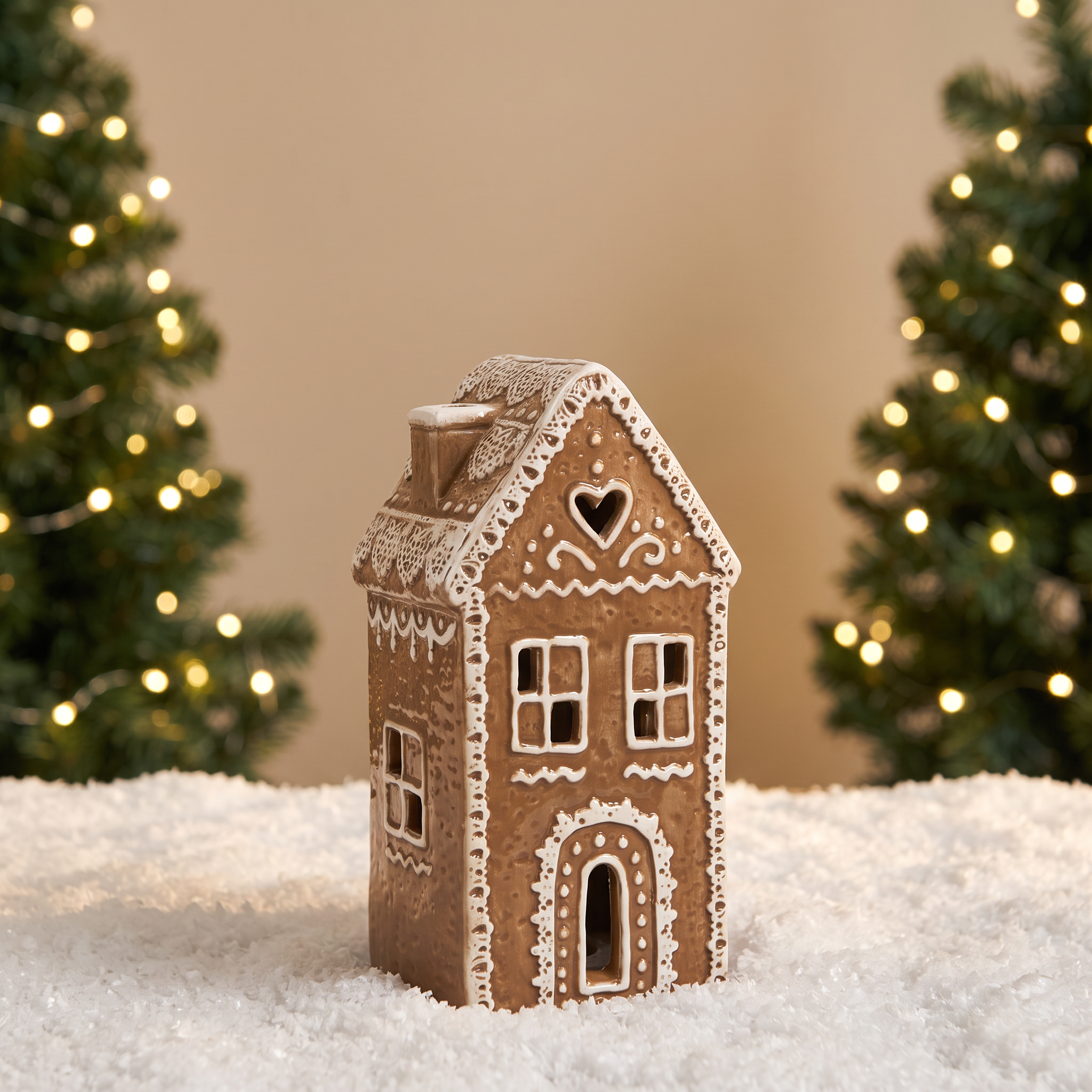 Figurine Chalet de Noël Ginger 16 cm en céramique effet pain d'épices, LED allumée, posée sur la neige devant un sapin lumineux flouté