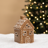 Figurine Chalet de Noël Ginger 12 cm en céramique effet pain d'épices, LED allumée, posée sur la neige devant un sapin lumineux flouté